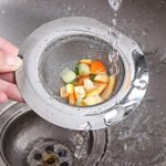 A hand holds a metal sink strainer containing small pieces of food. Water is being poured over it. The strainer is positioned above a stainless steel sink drain. | MySmartiee