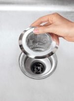 A hand is holding a metal mesh sink strainer above a kitchen sink drain. The strainer is positioned to be placed into or removed from the drain, which features a black circular component. The sink surface is stainless steel. | MySmartiee