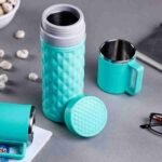 A textured turquoise Stainless Steel Insulated Refreshing Flask with its lid off and matching mug sit on a gray surface, surrounded by small white snacks, a notebook, and glasses, making it ideal for hot & cold drinks anytime. | MySmartiee